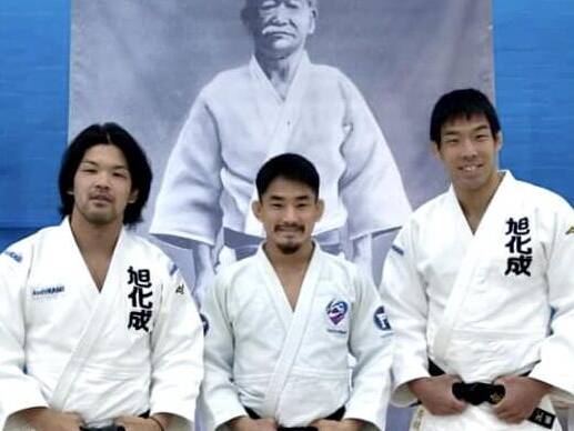 Ono, Nagasse and Take standing in their Judo Suit by Kano banner.