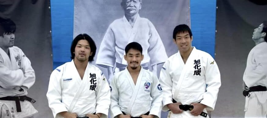 Ono, Nagasse and Take standing in their Judo Suit by Kano banner.