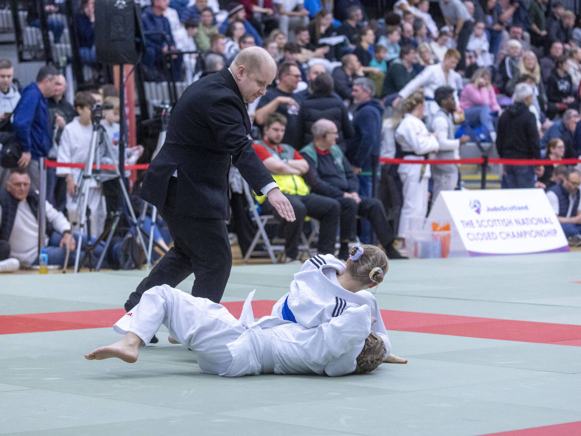 SNC action shot a referee stand on the mat as one judoka holds down another mid competition.