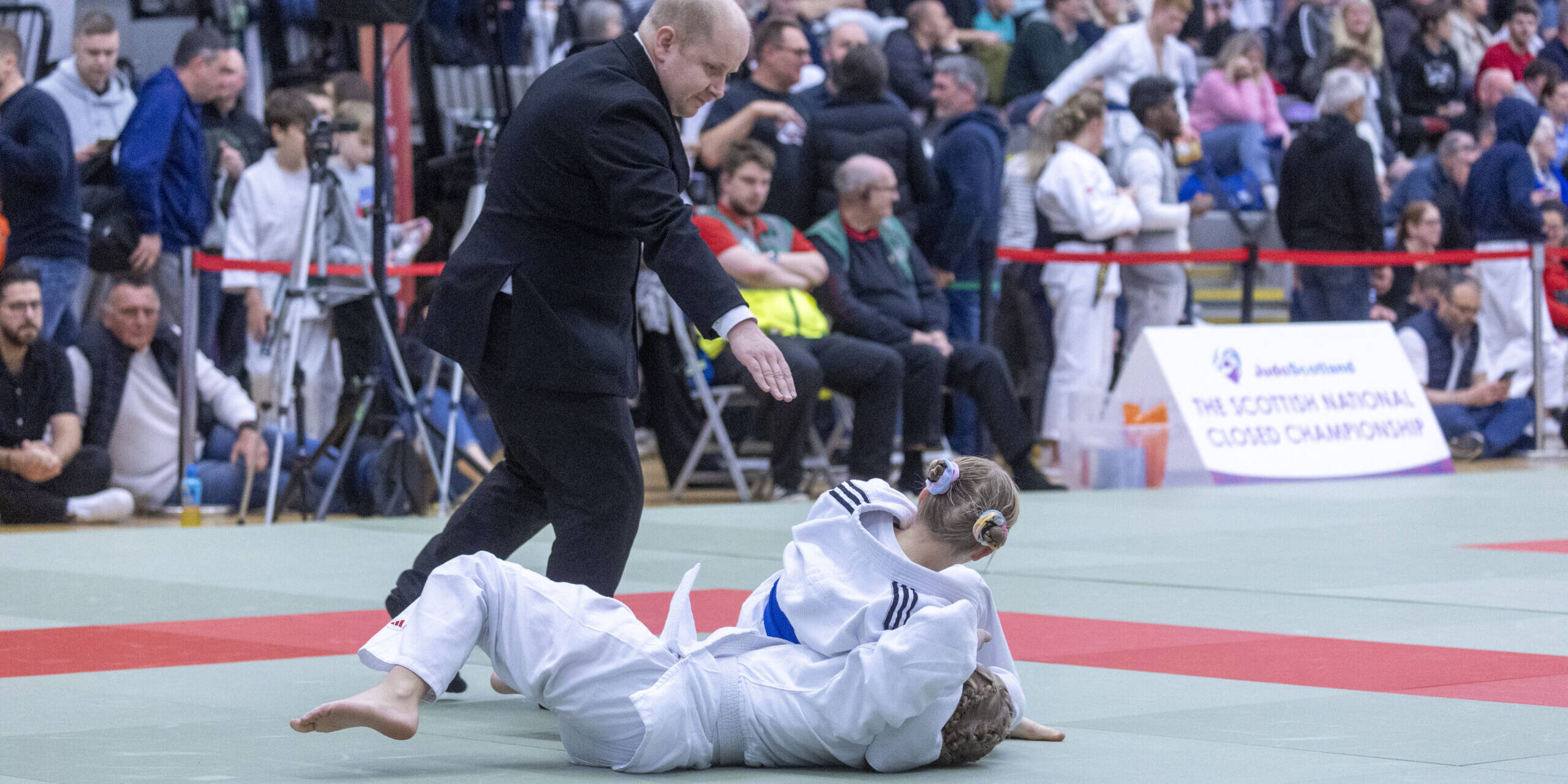 SNC action shot a referee stand on the mat as one judoka holds down another mid competition.